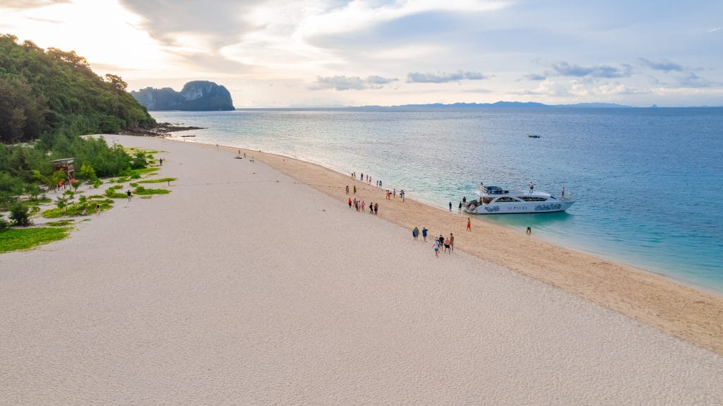 Long white sand spit with visitors and a tour boat moored in calm turquoise water on a dry-season day near Phuket