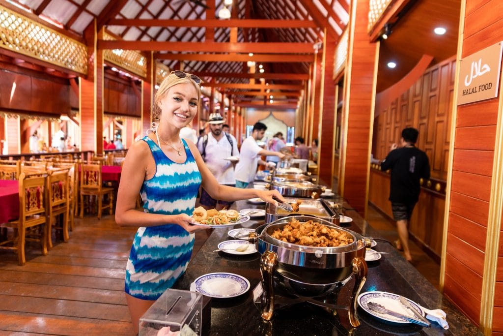 Smiling traveller serving herself from a Thai lunch buffet in a wooden restaurant with a halal food sign