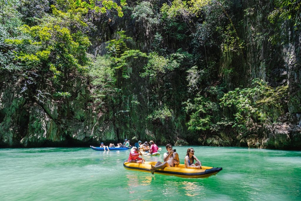 Visitors canoeing on calm green water beneath jungle-covered limestone cliffs in Phang Nga Bay