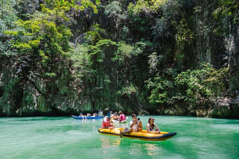 Visitors canoeing on calm green water beneath jungle-covered limestone cliffs in Phang Nga Bay
